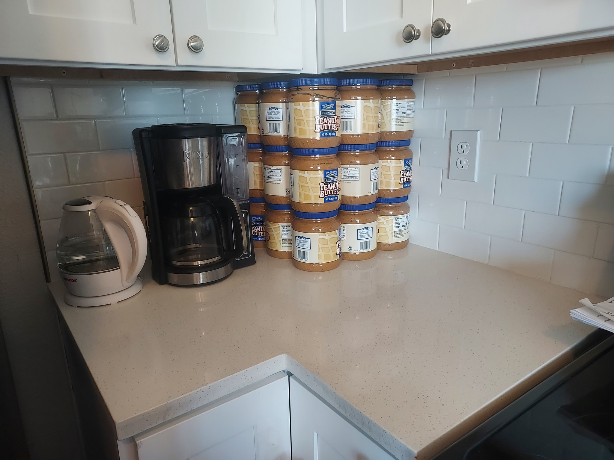 Peanut butter is love. Image depicts a kitchen counter with cabinets above. From left to right on the counter are a kettle, a coffee maker, and many jars of peanut butter. The jars appear to be arranged in a 3x3x3 cube, implying the presence of 27 jars in total.